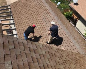 Aerial view of two roofing professionals inspecting a brown asphalt shingle roof on a residential home. The inspectors are walking across the roof, closely examining the shingles for signs of damage or wear, with neighboring homes visible in the background. Roof Inspection Arlington VA