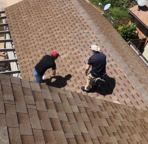 Aerial view of two roofing professionals inspecting a brown asphalt shingle roof on a residential home. The inspectors are walking across the roof, closely examining the shingles for signs of damage or wear, with neighboring homes visible in the background.