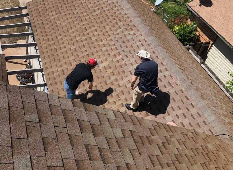 Aerial view of two roofing professionals inspecting a brown asphalt shingle roof on a residential home. The inspectors are walking across the roof, closely examining the shingles for signs of damage or wear, with neighboring homes visible in the background.
